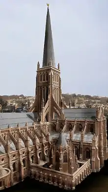 The central tower of a large gothic cathedral. The central tower is buttressed and with an imposing wooden spire. An octagonal chapter house is in the foreground.