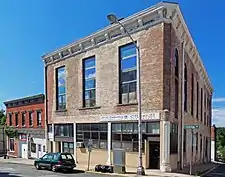 Two brick buildings seen from across the street and slightly to their left. The one closer to the camera has a higher second story with long tall windows. Its bricks have traces of a past white covering.