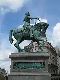 Photograph of a bronze statue of Joan of Arc on a horse, all on a large plinth. The picture is taken from below. She is in armour and carries a sword in her outstretched right hand. The horse has its head bowed and its right foreleg raised, as if trotting. The bronze statue is green from verdigris.