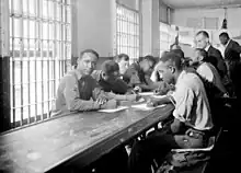 Black and white image of several prisoners, mostly of African heritage, sitting at a desk and writing. There are bars on the windows.