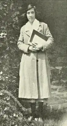 A young white woman, standing outdoors, wearing a coat and holding books and papers in her arms