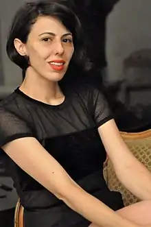 Ortal Ben Dayan, 3/4 angle, seated, wearing a black short-sleeved outfit. Dark hair in a bob hairstyle, and wearing orange-red lipstick.