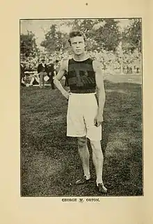 white man with short hair looking directly at the camera, one hand on hip and the other arm hanging straight down, wearing baggy white athletic shorts and a dark sleeveless jersey with a large "P" on the chest