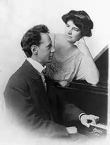 Ossip and Clara (in her forties) at a piano showing the upper half of their bodies. Ossip sits at the piano looks to the right with one hand resting on the keyboard. Clara is standing and leans her elbow on the top of the piano and looks at Ossip. Her hair is dark short and styled in waves.