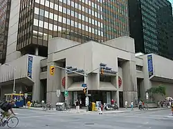 A block-shaped concrete building sits on a city street corner in front of a steel-and-glass skyscraper