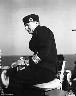 A man in naval uniform sitting on a seat on the bridge of a ship. He is wearing thick gloves and a cap, and is slightly side on to the camera. A body of water can be seen in the background.