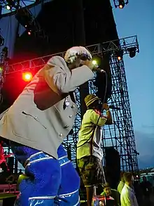 Two men rap into microphones whilst on stage. One wears a blonde wig, a grey jacket and blue trousers, and the other wears a green chequered hat, white shirt and khaki shorts.