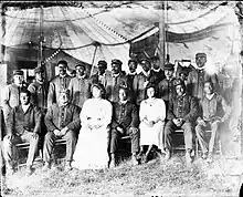 18 people (16 men and 2 women) sit under a big tent. The men wear uniforms and the women wear white dresses.