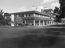 black and white image of a modernist building, two stories at the front, four stories at the back with trees and roads around it