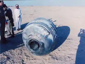 Cylindrical rocket fragment on sand, with men looking at it