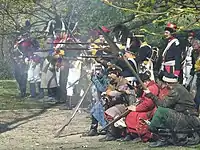 A group of reenactors dressed in Polish 18th century uniforms and szlachta civilian attire firing a musket volley.