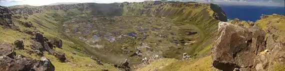 Colour photo of circular pond strewn marsh surrounded by steep cliff topped slopes – the sea is visible through a gap in the cliffs on the right