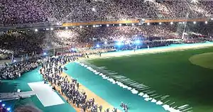 A portion of the stadium with stands full of people, a large artificial tree is on the right side of the image. A group of people are walking together on the stadium floor