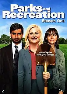 The DVD cover shows three people standing side-by-side. In the middle, a blond woman wears a gray dress suit, smiling and holding a golden shovel. On the right, a brown-haired woman in a green jacket looks at her and smiles. On the left, a black-haired man with a beard, wearing a gray suit and green tie, looks at the middle figure while smirking. Above the trio is the text, "Parks and Recreation Season One".