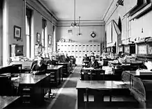 Black and white photo of eight men dressed in suits, sitting at large wooden desks in a news room