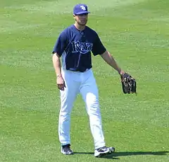 A man standing on a grassy surface wearing a baseball uniform with a dark blue shirt and white pants