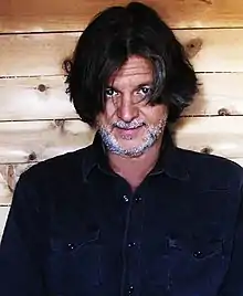 A man in front of a wooden panel wall, with black hair parted from the middle, wearing a black shirt.