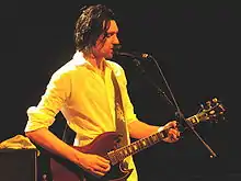 A thirty-year-old man is playing a six-string electric guitar while singing into a microphone.