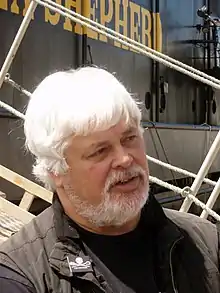 A heavyset man with white hair and a white beard, standing in front of a boat with the words "Shepherd" written on it in yellow paint