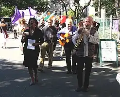 A line of people performing various instruments outdoors; monochromatic flags of various colors are seen in the background