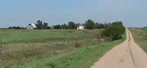 Dirt road running through gently rolling grassland; house and barn on near horizon