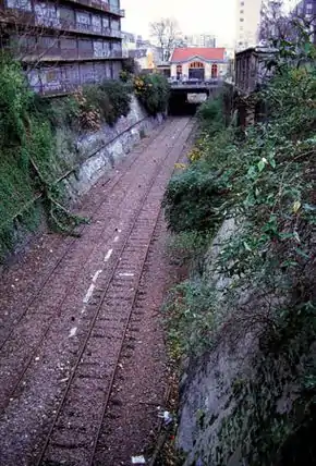 Below-grade railroad track, with small building behind a bridge above it in background