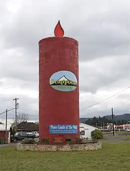 A cylindrical-shaped red tower-like structure resembling a candle sits inside a circular stone brick wall with plants and shrubs around it. A red flame-like structure sits atop the candle, and an oval-shaped logo with the word "Scappoose" in front of a drawing of a mountain rests in the center of the candle. A blue banner with the words "Peace Candle of the World" in white is draped near the bottom of the structure. In the background is a rural setting with houses and trucks below a sky with gray clouds.