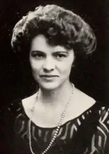 A white woman with dark hair in a bouffant style, wearing a dark dress with a scooped neckline and a strand of pearls