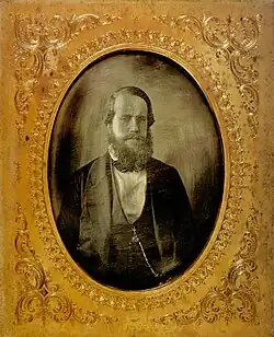 a gold-framed photographic portrait of a bearded man dressed in a dark formal suit with a watch chain hanging from the vest