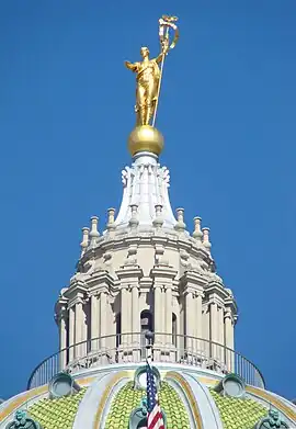 A stone, columned cupola atop a green, tiled-dome. The cupola is topped with a gold globe, upon which stands a gold statue with an outstretched arm and holding a staff.