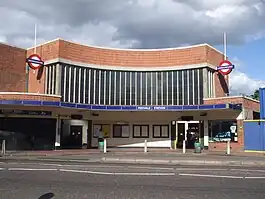 A red-bricked building with a rectangular, dark blue sign reading "PERIVALE STATION" in white letters all under a light blue sky