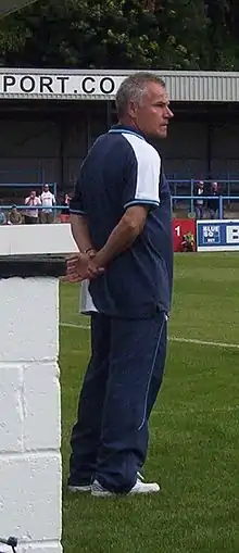 A middle-aged man with greying hair, wearing a dark blue T-shirt and matching jogging bottoms, standing near the touchline of a sports pitch.  Some spectators are visible in the background.