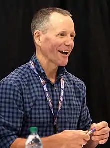 A three-quarters bust portrait photo shows a middle-aged Caucasoid man looking to the photographer's right; he is wearing a blue-plaid collared shirt and a lanyard.