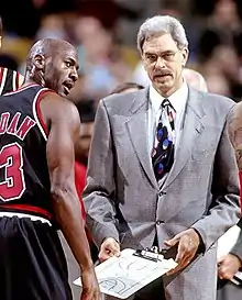 Man in gray hair, glasses, and suit on the edge of a basketball court and beside a player in uniform