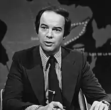 Black and white photo of Nadeau in a suit and tie behind a desk on a television news set