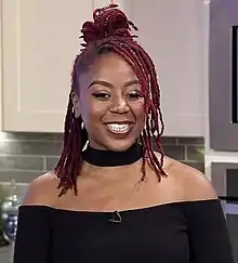 The woman, Pinky Cole, has brown skin and dyed red locs and is smiling at the camera.