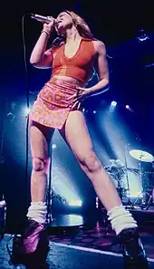 A woman in an orange top with a short pink skirt, white socks, and black trainers with heart patterns singing at a microphone on a stage.