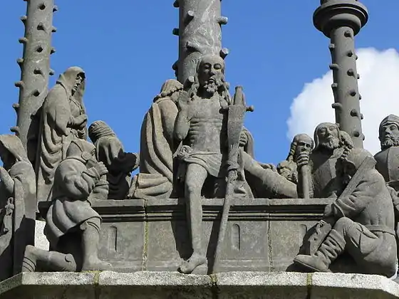 Jesus steps from his tomb in the scene at Plougonven depicting the resurrection. He gives a blessing with his right hand and he carries a cross in his left hand. Beside the tomb two soldiers sit sleeping.