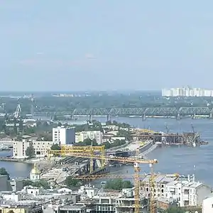 Southern end of the Rybalskyi Peninsula, pictured from the Podil. Kyiv Harbour entrance and Podilskyi Metro Bridge (under construction) are seen on the foreground.