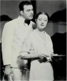 white man in waiter's uniform standing intimately close behind a bespectacled woman