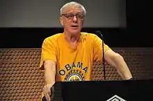 A gray-haired man with glasses wearing a yellow shirt standing at a podium