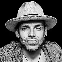 black-and-white picture of man with contour facial hair and two earrings, wearing a brimmed hat, smirking at camera