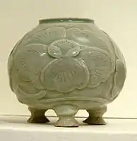 A blue-green tinted grey pot with a wide bottom supported by three stubby legs, a wide body, and a smaller opening at the top. A large five petal flower pattern is out-dented on the body of the jar.