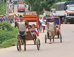 Two Malagasy men pull children in rickshaws. Both modern vehicles and more rickshaws can be seen in the background.