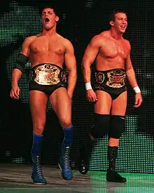 Two dark-haired men are both wearing black wrestling tights. One wears knee-high blue wrestling boots with blue knee-pads and a black elbow pad on his right arm, and the other wears black knee-high wrestling boots with black knee-pads, and white tape around his wrists. The two are wearing professional wrestling championship belts around their waists.