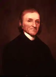 Half-length portrait of an elderly man with wispy brown hair. He is wearing a black jacket and a white shirt.