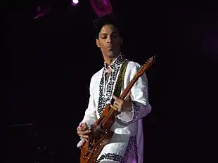 Prince playing the guitar wearing a white, jeweled shirt.