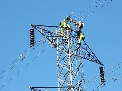 Cable riggers atop a pylon adding a fiber optic data cable wound around the top tower stay cable. The cable (SkyWrap) is wound on by a traveling machine, which rotates a cable drum around the support cable as it goes. This travels under its own power from tower to tower, where it is dismantled and hoisted across to the opposite side. In the picture, the motor unit has been moved across but the cable drum is still on the arrival side.