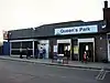 A brown-bricked building with a white sign reading "Queen's Park" in blue letters underscored by a green line and a blue line