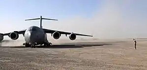 Colour photo of a grey military aircraft on a dusty airstrip. A person is standing to the right of the aircraft.